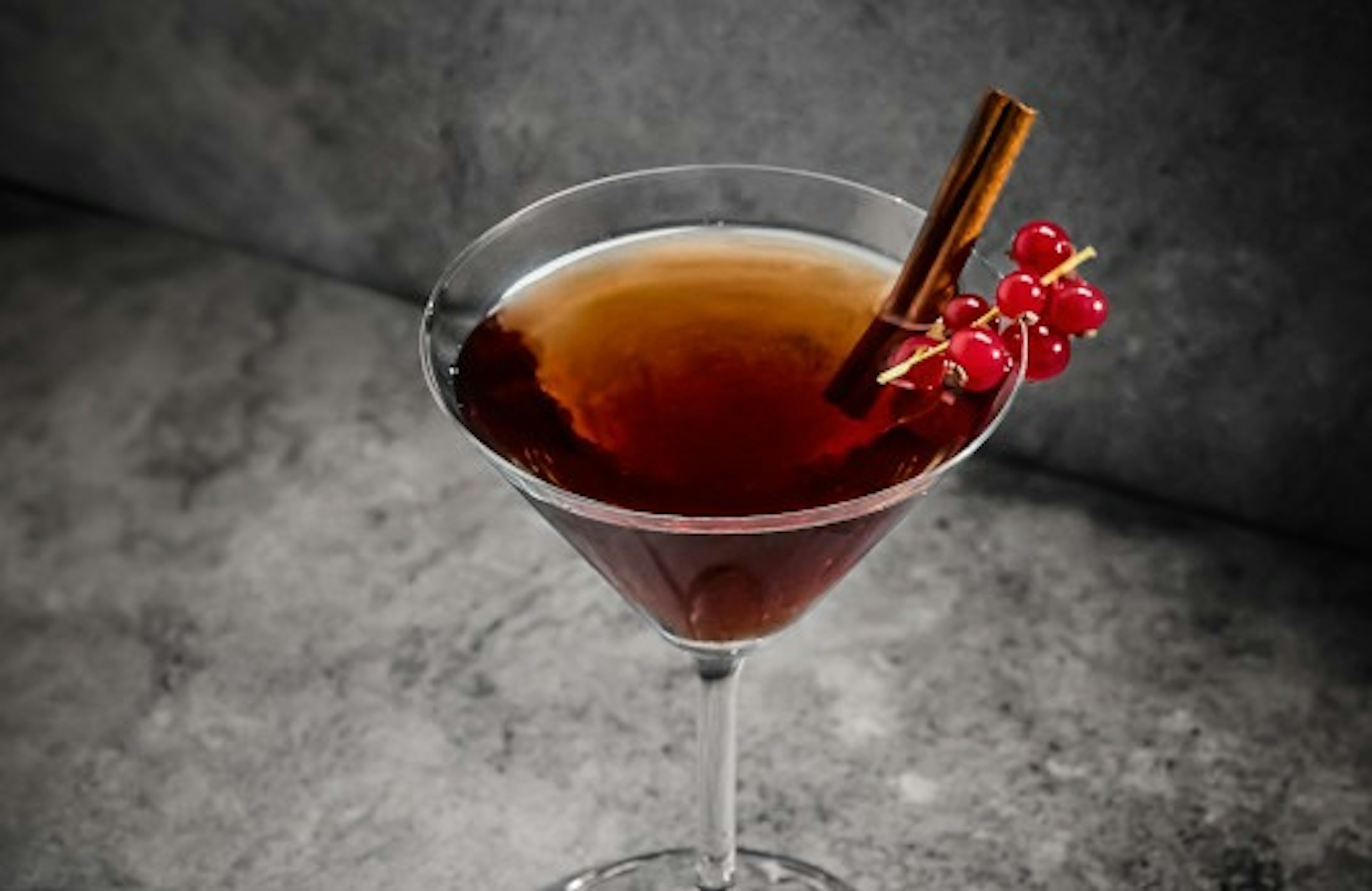 Um cocktail escuro num copo de martini, com um pau de canela e groselhas vermelhas, sobre um fundo cinzento escuro