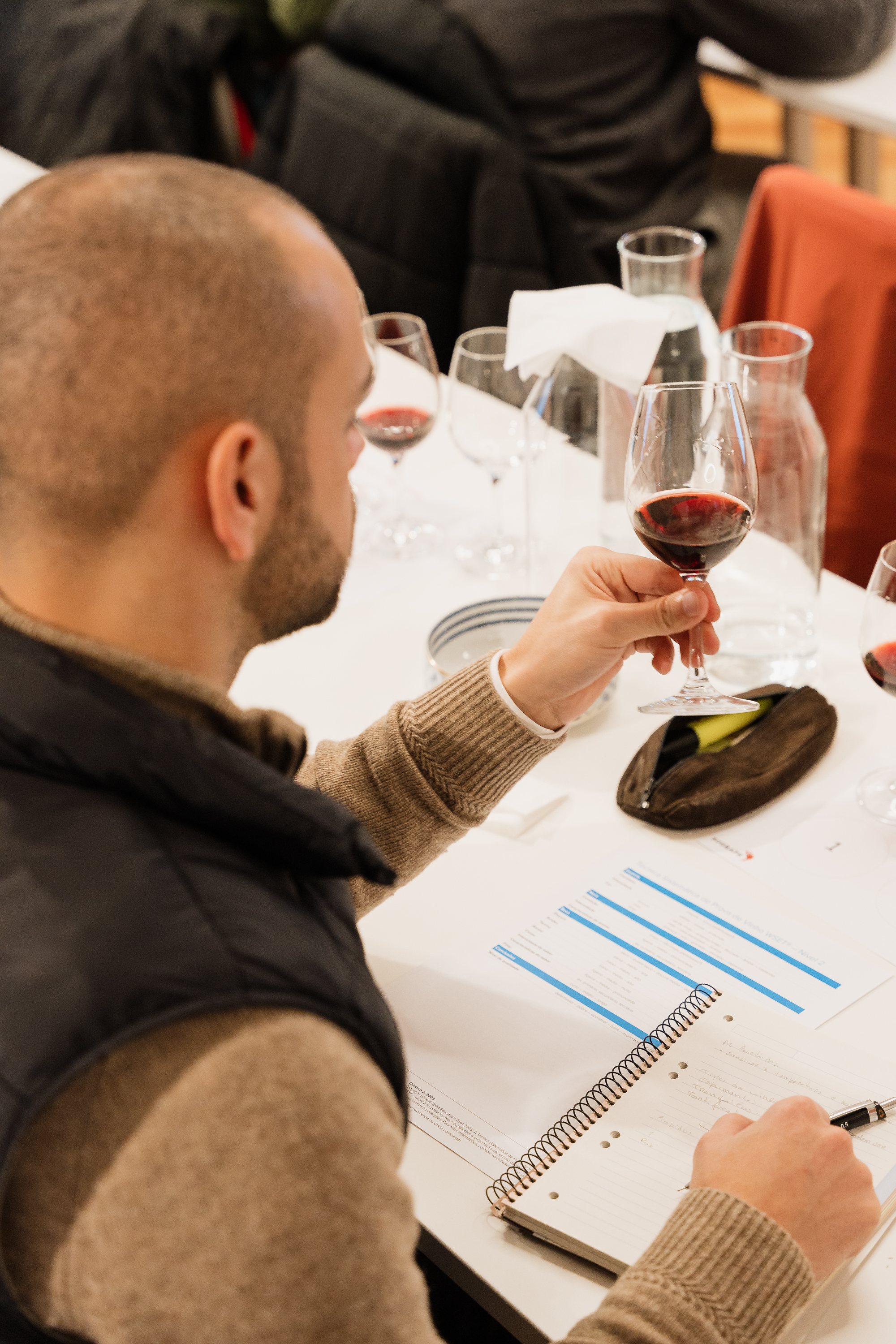 Um homem está a segurar um copo de vinho tinto numa prova ou aula, enquanto toma notas