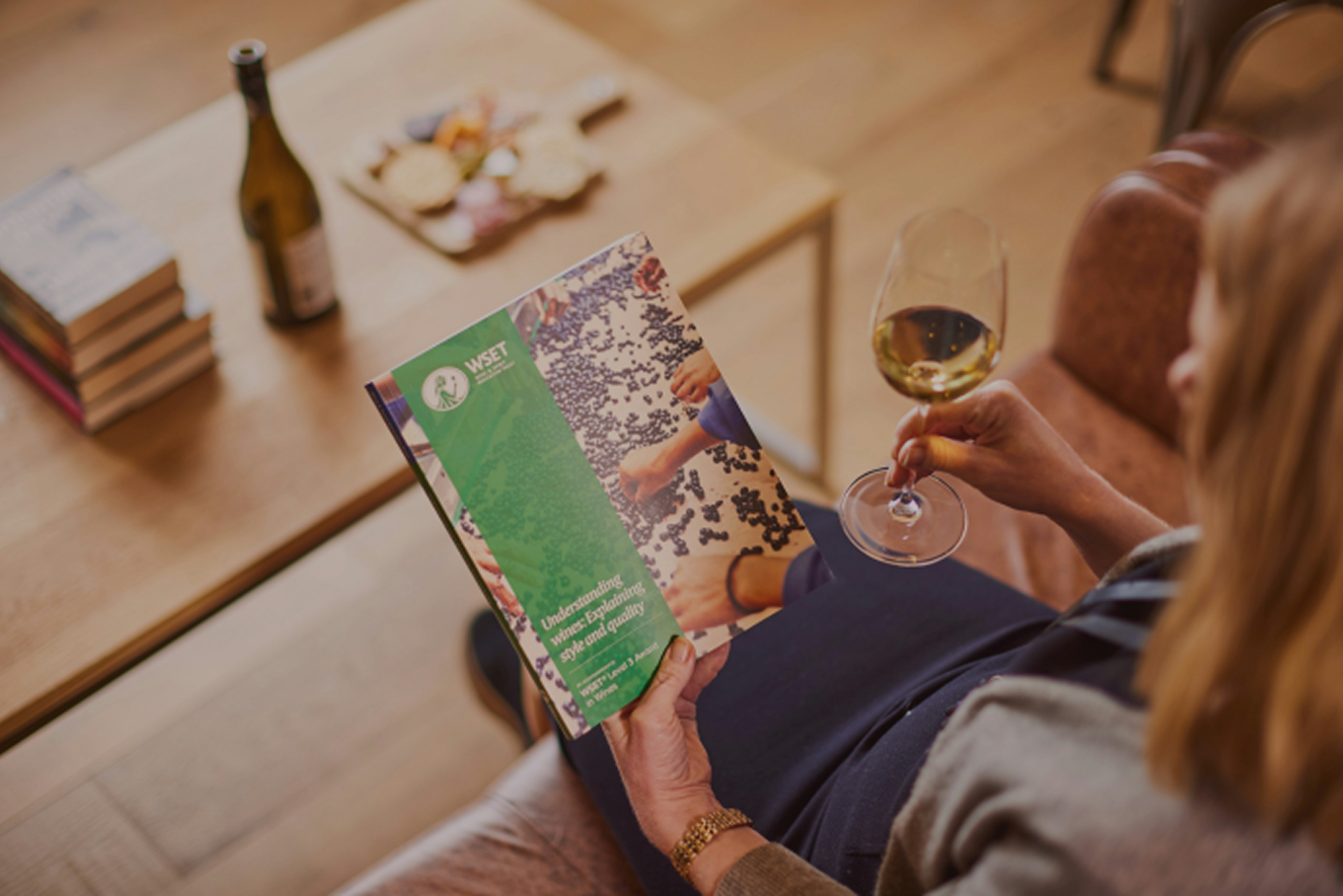 Uma pessoa segura um copo de vinho e lê uma publicação sobre vinho, enquanto relaxa numa sala com uma garrafa de vinho e aperitivos na mesa