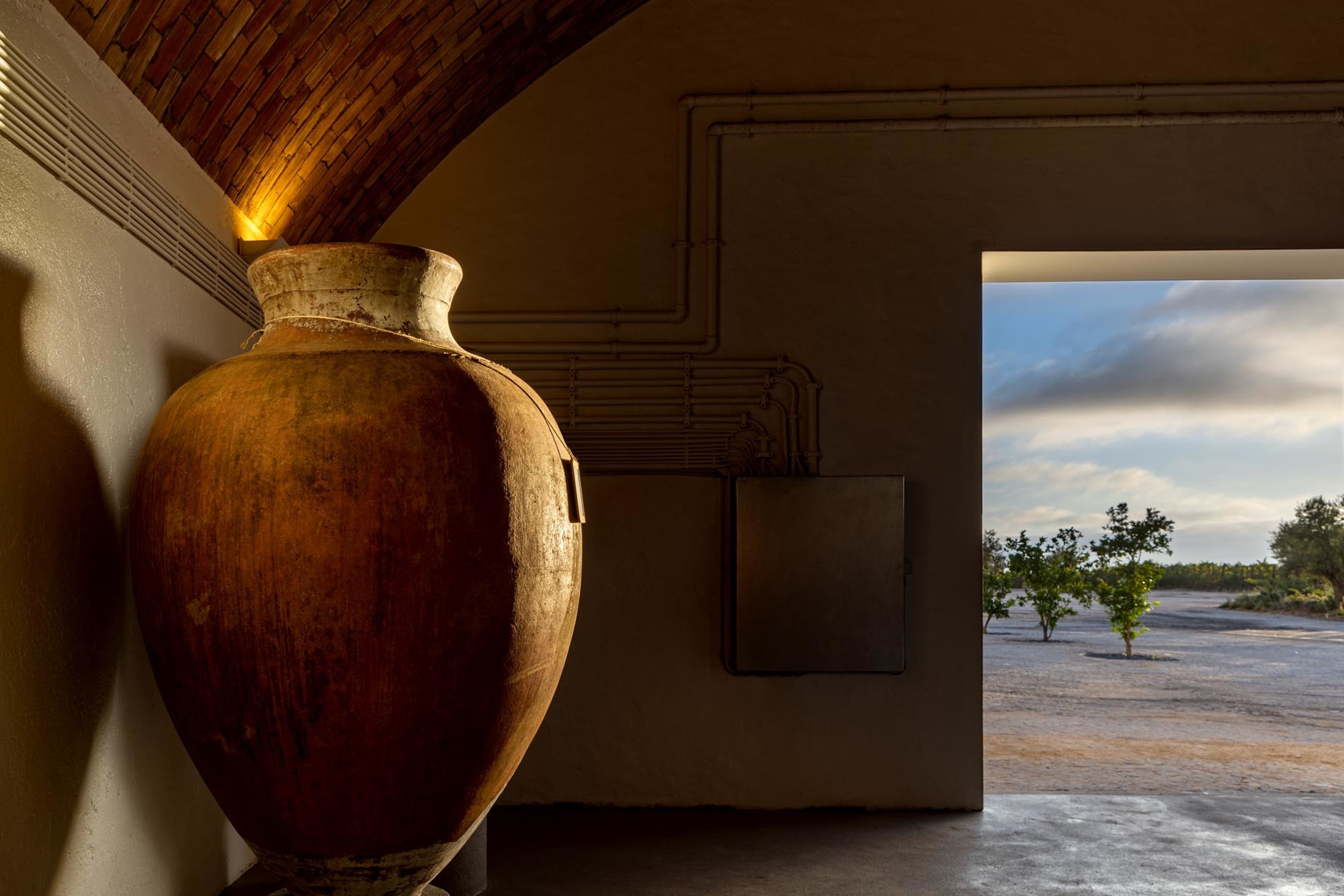 Uma grande ânfora de barro iluminada dentro de um edifício, com uma abertura que revela uma paisagem exterior