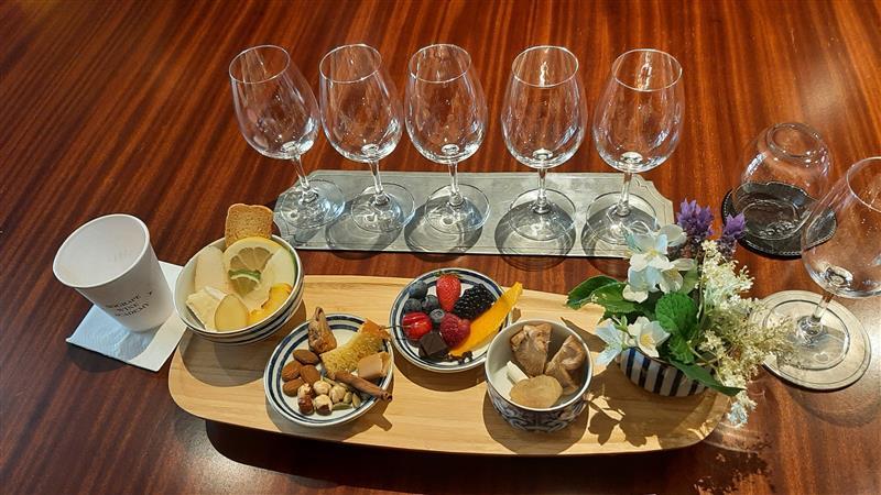 Uma mesa de madeira apresenta vários copos de vinho vazios, um tabuleiro com diversos petiscos, frutas e nozes, e um pequeno arranjo floral