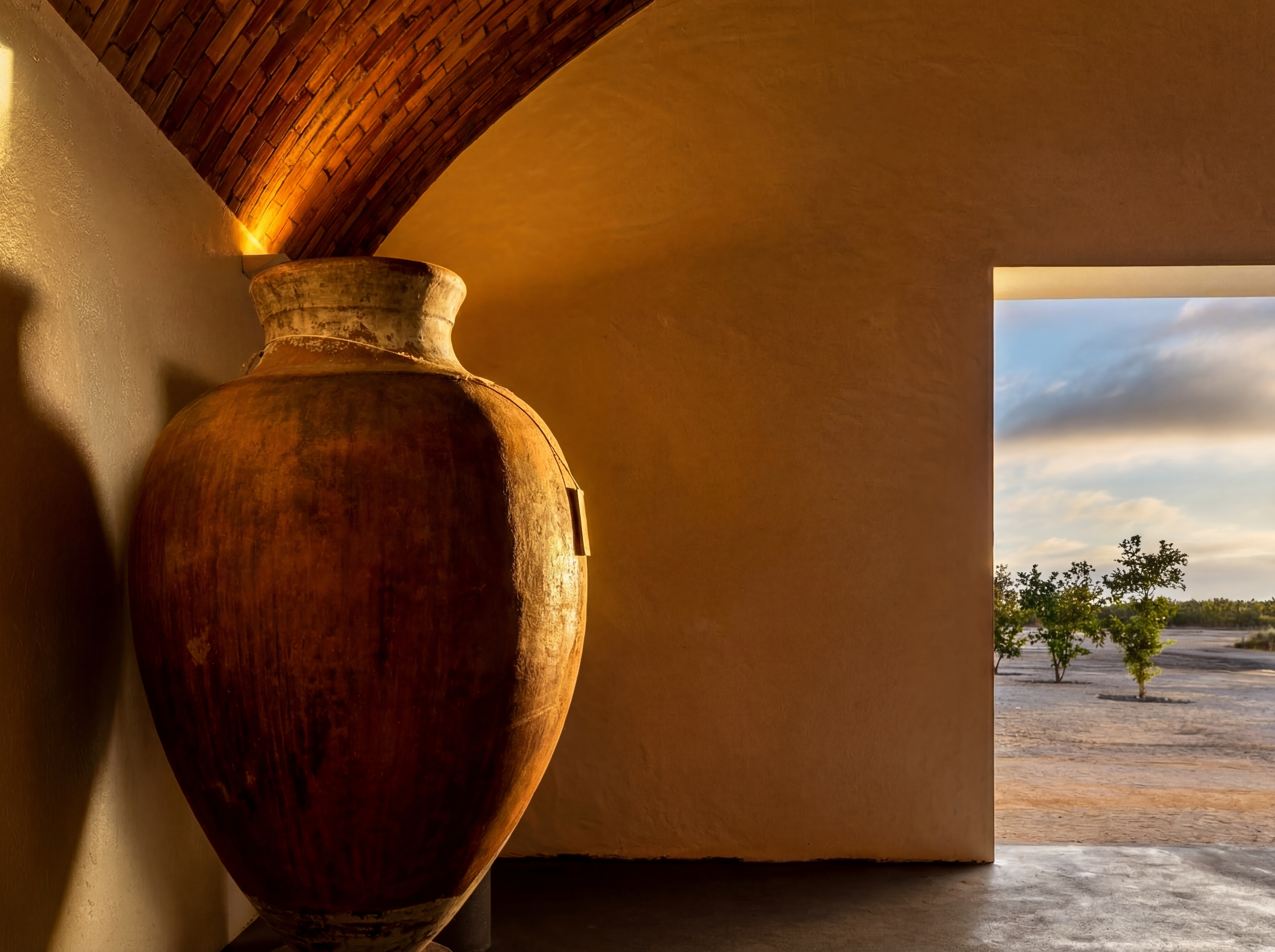 Uma grande talha de barro é iluminada por um teto abobadado de tijolos, com uma abertura para uma paisagem exterior de árvores esparsas e céu