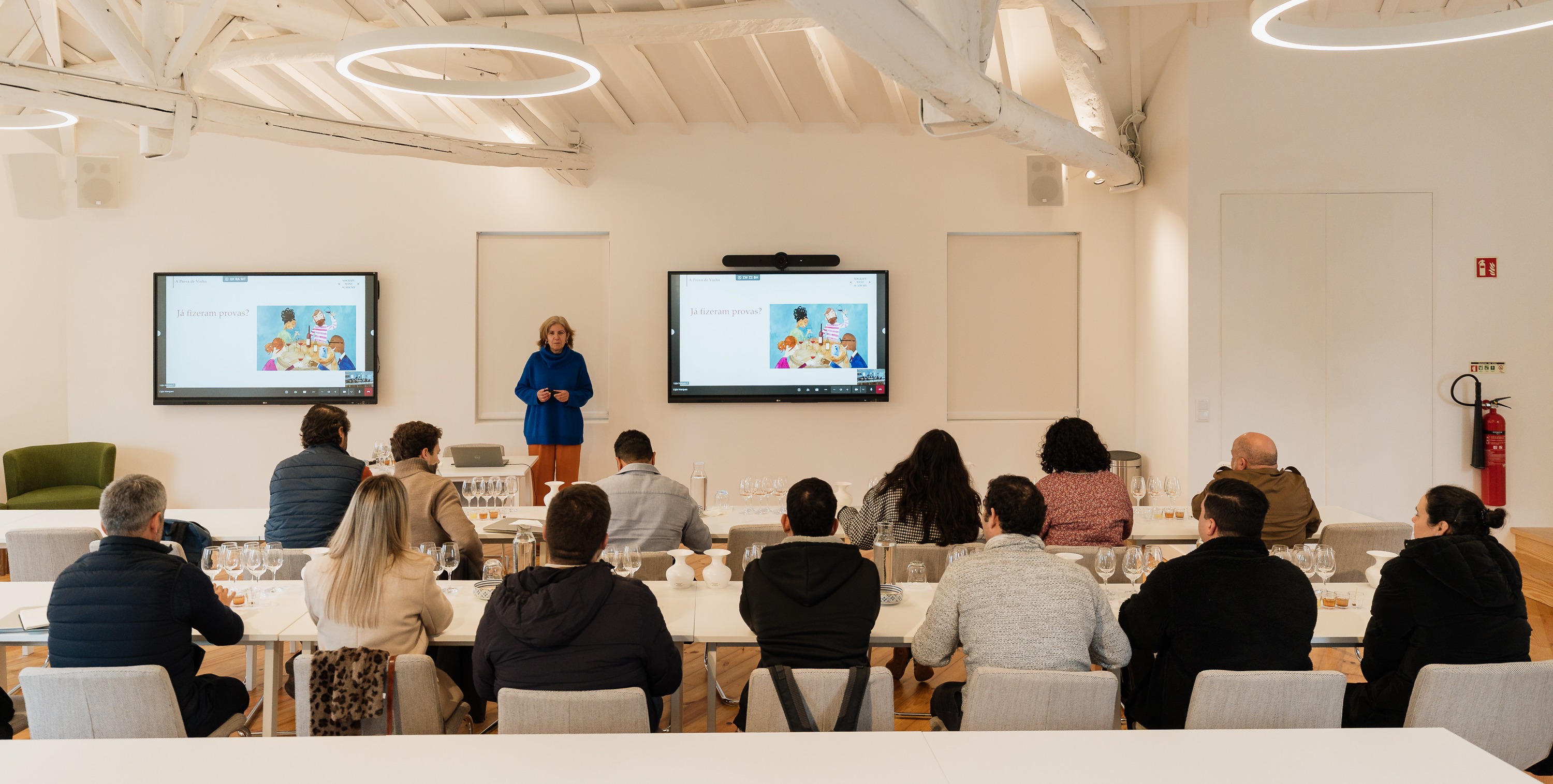 Uma oradora faz uma apresentação em frente a dois ecrãs a um grupo de pessoas sentadas em longas mesas com copos para degustação, numa sala de formação iluminada