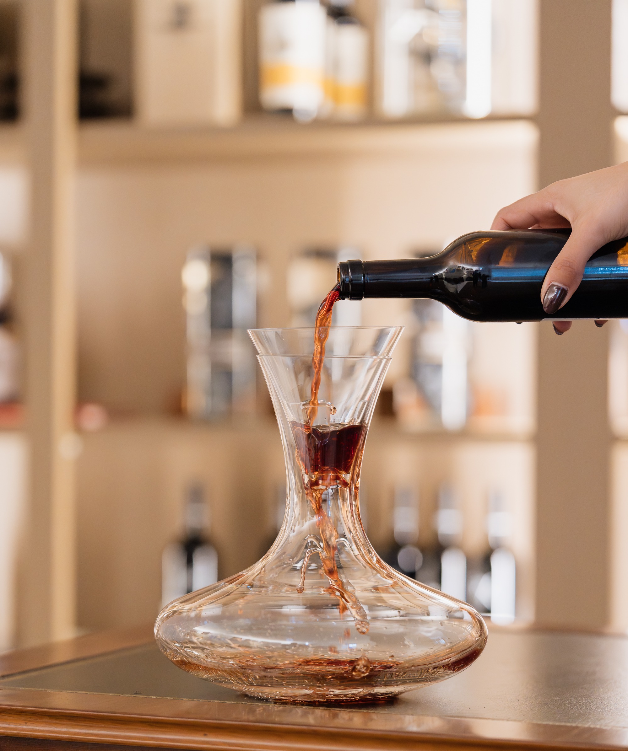 Uma mão verte vinho tinto de uma garrafa para uma garrafeira de vidro, sobre um móvel de madeira, com garrafas em prateleiras ao fundo