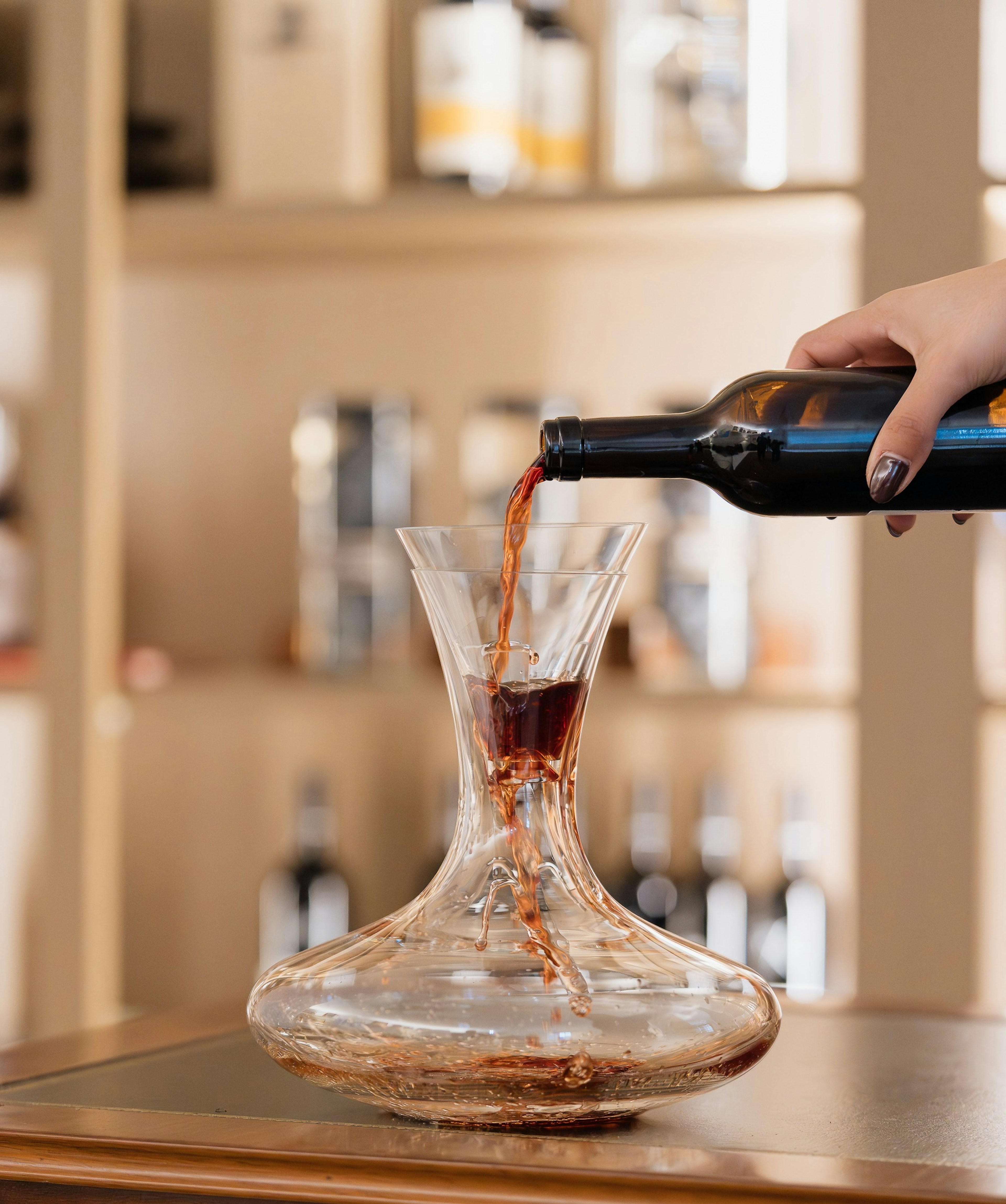 Uma mão verte vinho tinto de uma garrafa para uma garrafeira de vidro, sobre um móvel de madeira, com garrafas em prateleiras ao fundo