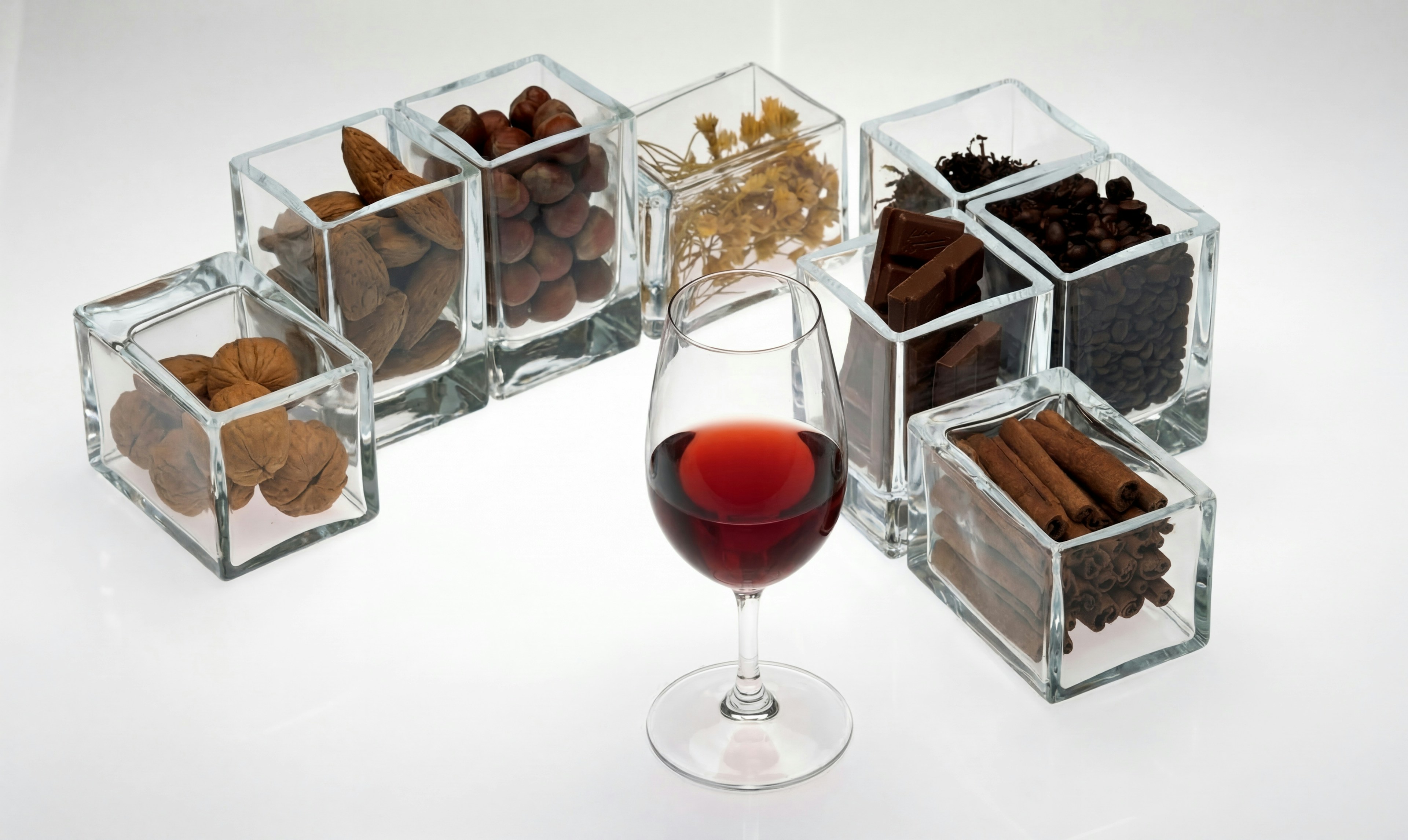A imagem mostra um copo de vinho tinto rodeado por pequenos recipientes de vidro com frutos secos, ervas, café, chocolate e canela