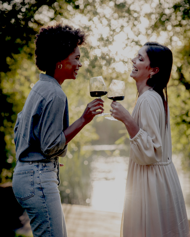 Duas mulheres sorridentes brindam com vinho tinto ao ar livre, junto a um lago