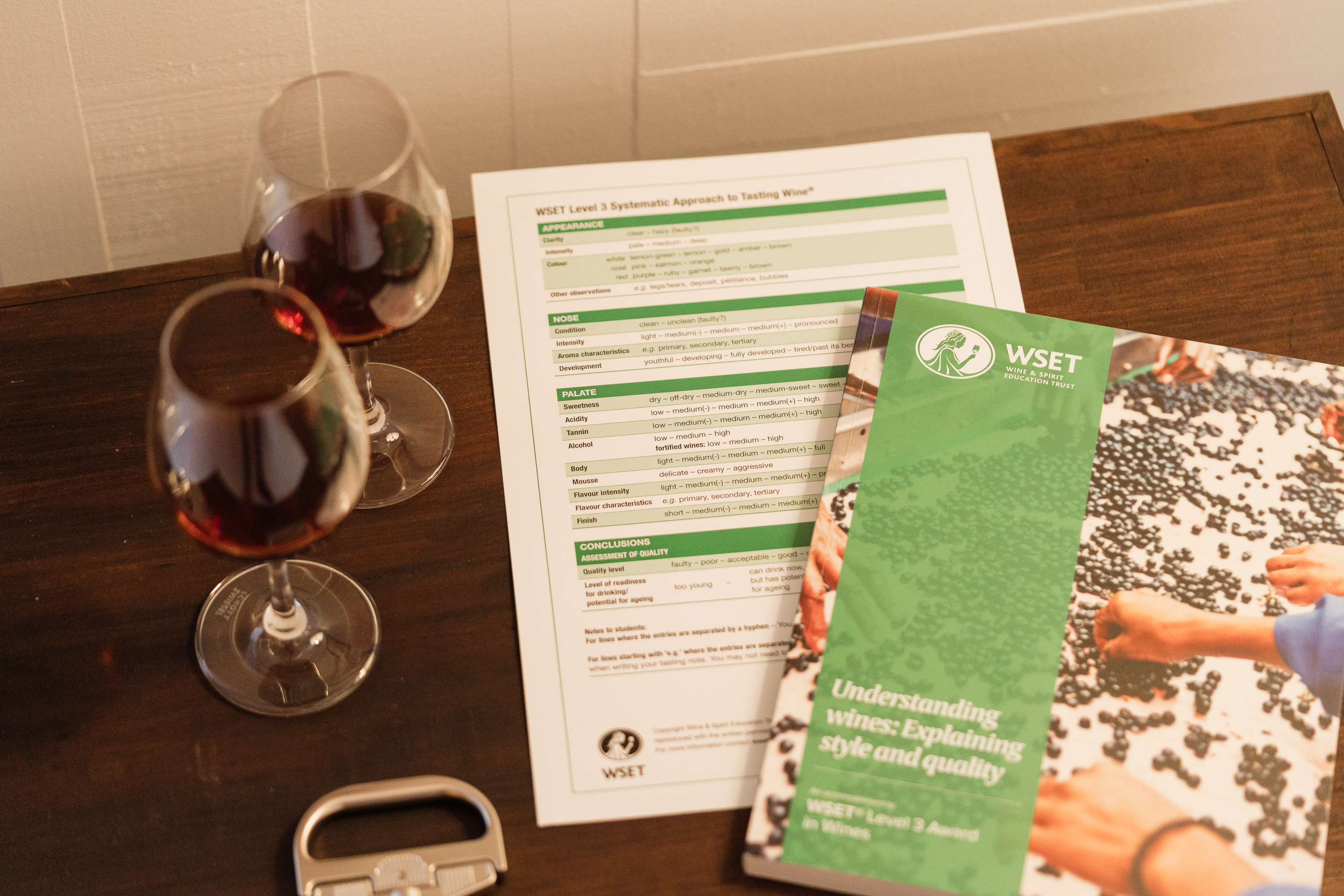 Dois copos de vinho tinto, uma ficha de prova WSET e um folheto sobre vinhos numa mesa de madeira