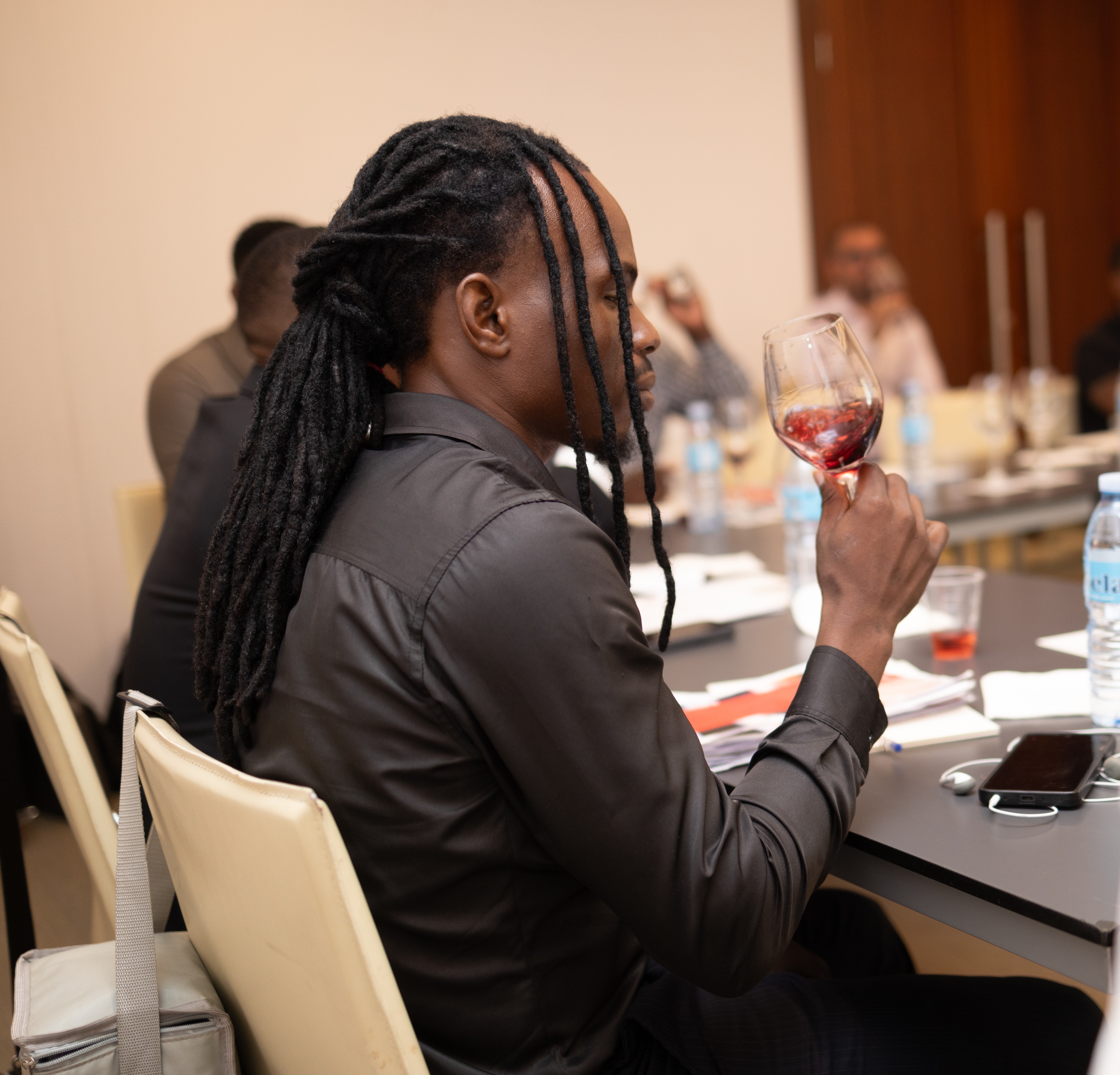 Um homem de origem africana com dreadlocks está sentado a uma mesa, segurando um copo de vinho tinto, num ambiente de reunião
