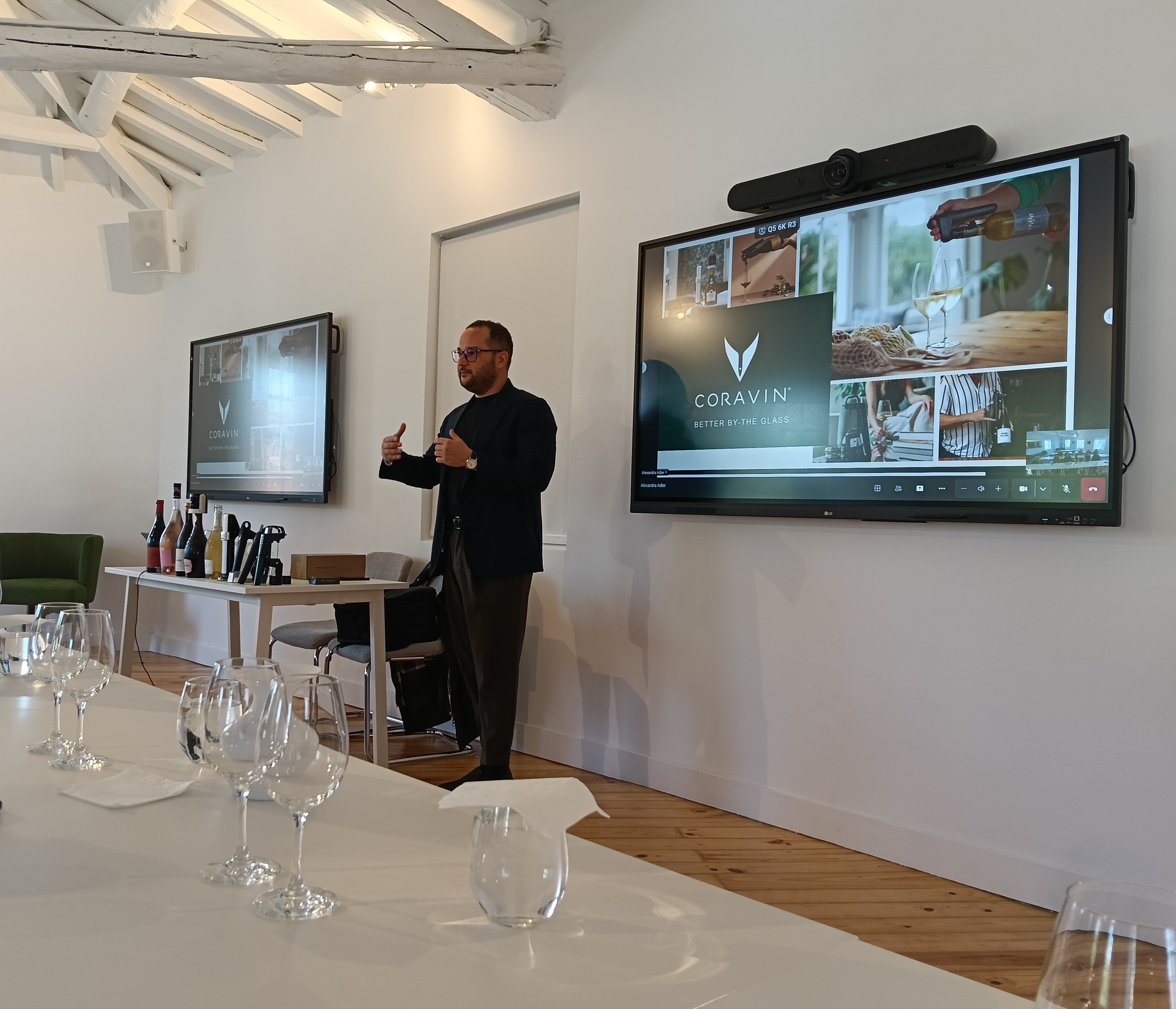 homem de óculos gesticula com os polegares para cima enquanto apresenta num evento com dois grandes ecrãs a exibir a marca "Coravin", garrafas de vinho e copos