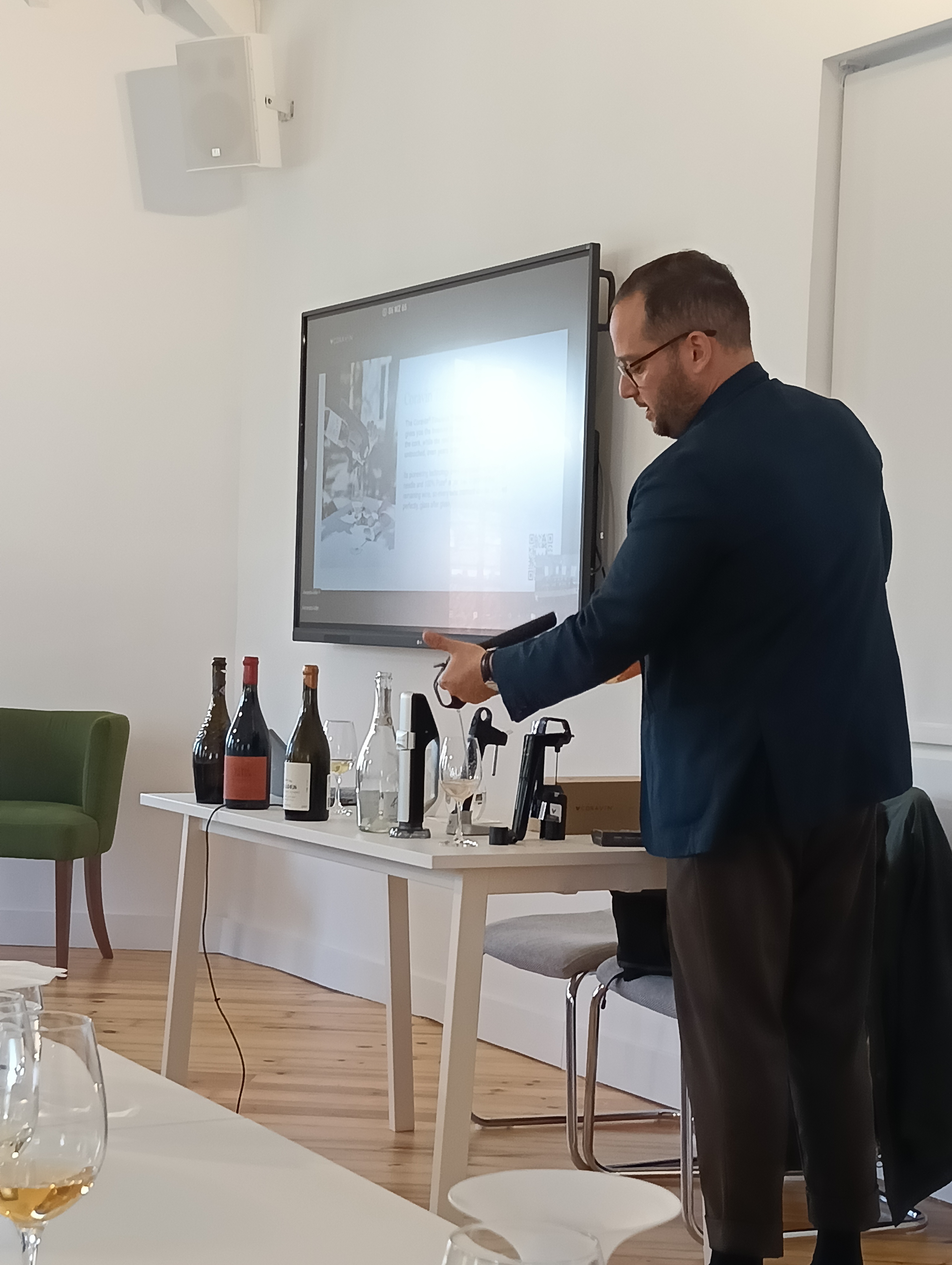 O mesmo homem, de perfil, inclina-se sobre uma mesa com garrafas de vinho e dispositivos Coravin, apontando ou interagindo com os objetos, enquanto um ecrã exibe conteúdo atrás dele