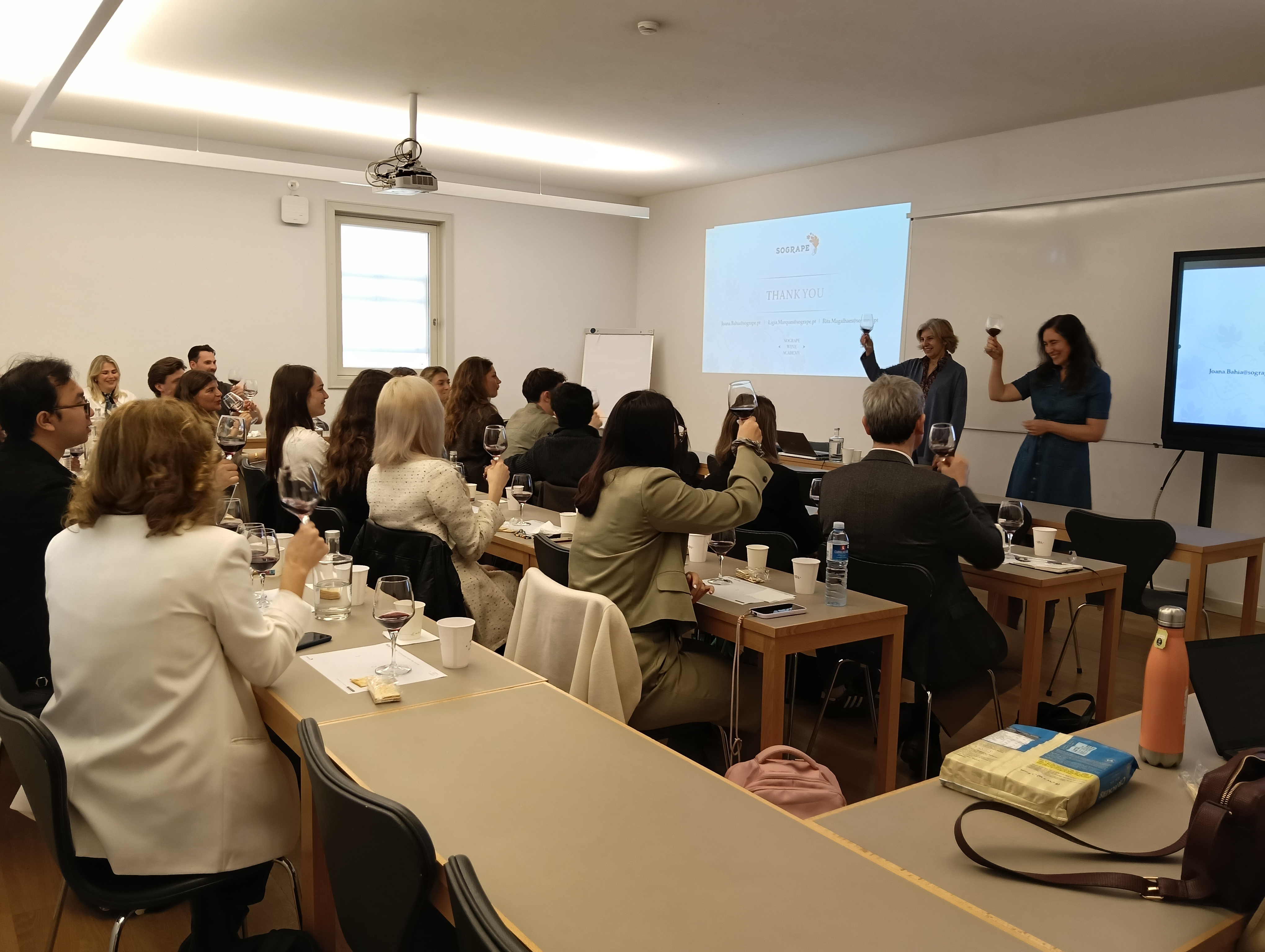 Pessoas estão sentadas em mesas de sala de aula, muitas delas com copos de vinho tinto, enquanto duas mulheres à frente levantam os seus próprios copos diante de um ecrã com a palavra "THANK YOU"