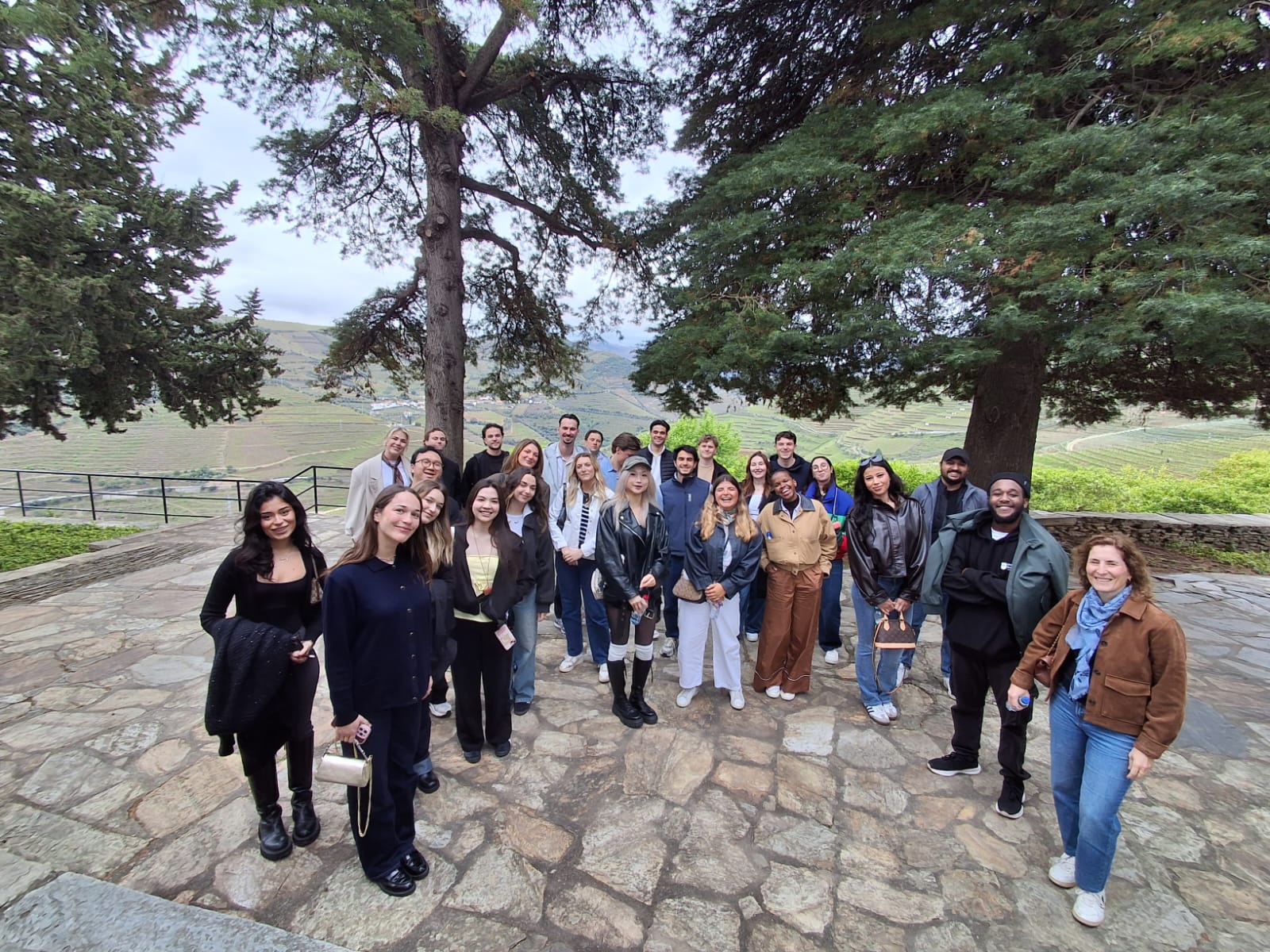 Um grupo numeroso de pessoas posa para uma fotografia num pátio de pedra, com duas grandes árvores a emoldurar um vale de vinhas em socalcos e um céu nublado ao fundo
