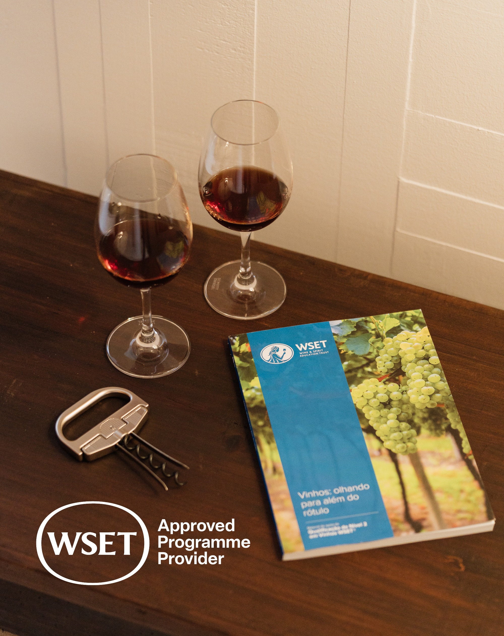 imagem com 2 copos de vinho com vinho tinto dentro, 1 saca rolhas e um livro WSET sobre mesa castanha escura com selo/logo WSET approved provider
