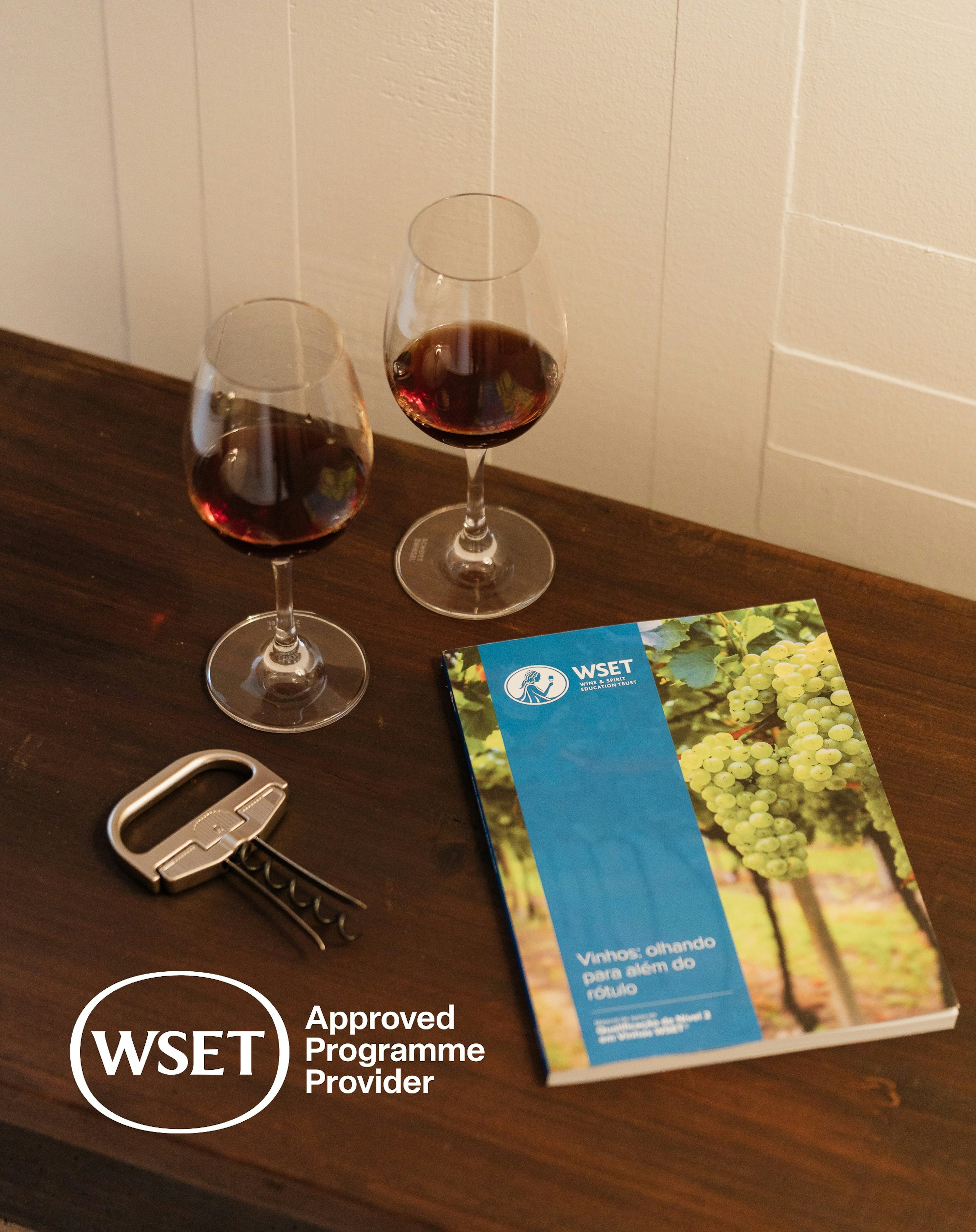 imagem com 2 copos de vinho com vinho tinto dentro, 1 saca rolhas e um livro WSET sobre mesa castanha escura com selo/logo WSET approved provider