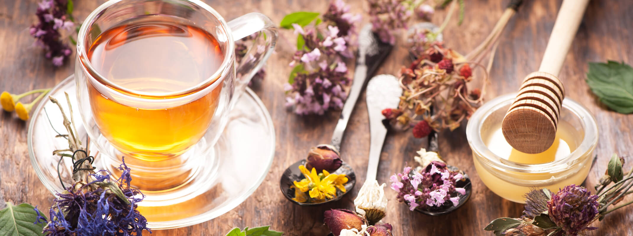 Clear glass of herbal tea surrounded by fresh herbs and honey next an image of a woman sitting looking towards a window and enjoying a cup of tea that is sitting on the table