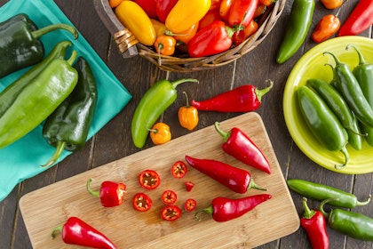 A variety of sweet and hot peppers in a basket, on a napkin, on a cutting board and on a plate - all on a wooden table