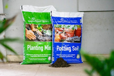 2 bags of soils - SummerWinds Nursery's Organic Planting Mix and Potting Soil with blurred plants in the foreground and a small pile of soil in front of the bags with a small plant in it