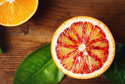 Half of a blood orange on a wood taben with plant leaves and half of a regular orange in the left corner