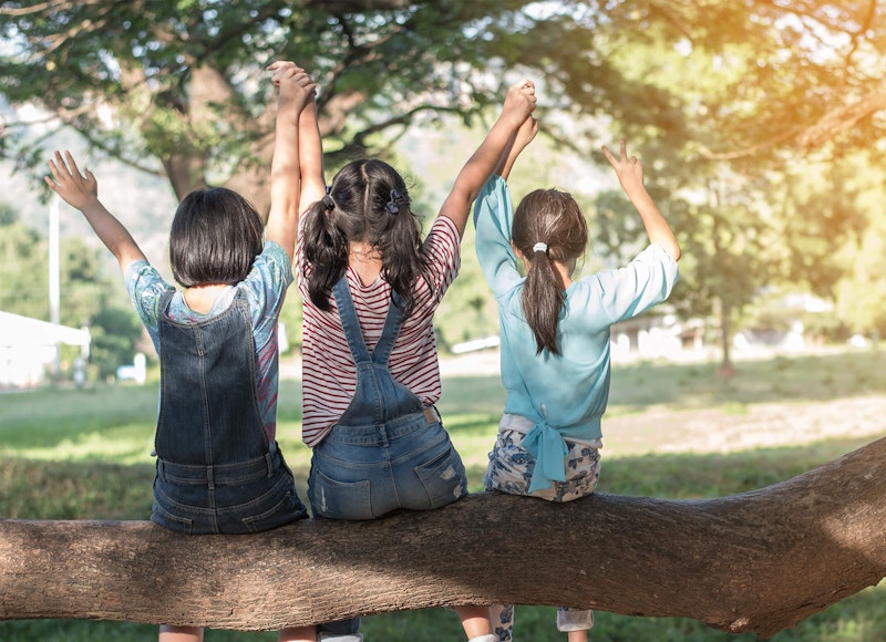 Three girls sitting on a large branch of a tree underneath its shade in a park