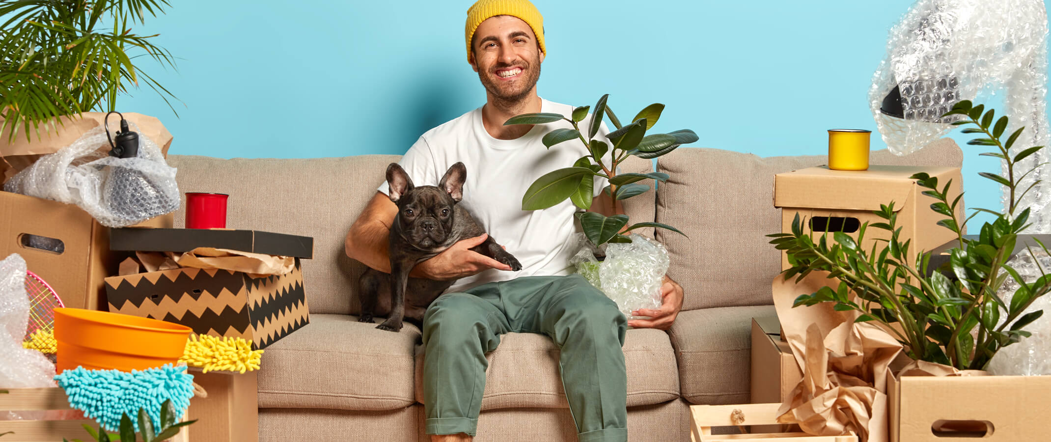 Man sitting on his couch with dog in one hand and rubber tree plant in the other smiling while in the midst of his things all still in a packed up state