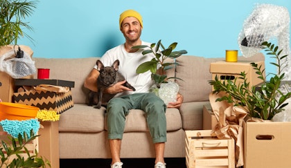 Man sitting on his couch with dog in one hand and rubber tree plant in the other smiling while in the midst of his things all still in a packed up state