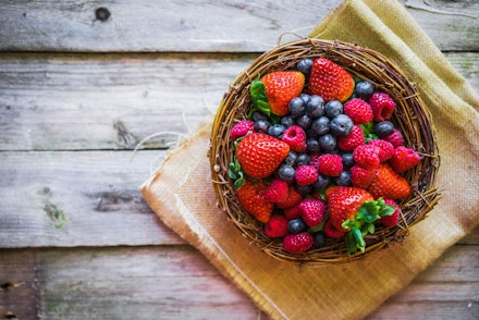 A bowl made out of twigs, filled with fresh strawberries, blueberries and raspberries on a piece of fabric, on top of a wooden table