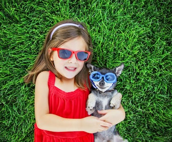 A girl in a red dress and sunglasses hugging her dog that's wearing blue sunglasses, both laying on a lush lawn