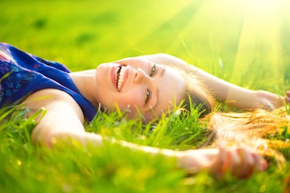 A smiling woman laying in the grass, wearing a blue shirt, with sun rays in the background