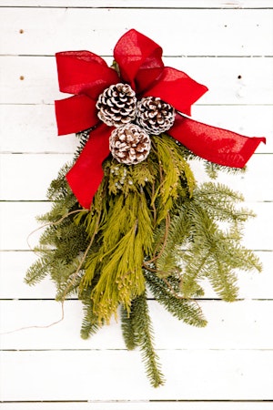 A close up a door charm made from fresh cut holiday greens with a red ribbon and pine cones