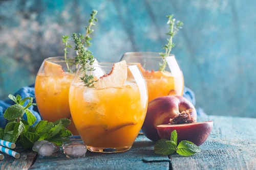 3 peach bellezza cocktails garnished with peaches and thyme surrounded with the same garnishes on a blue table