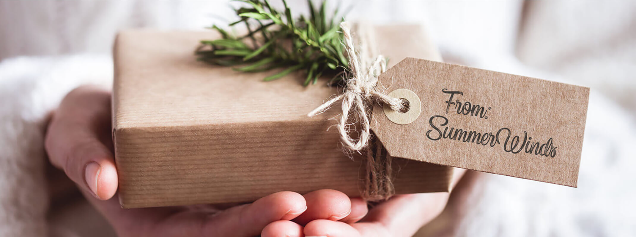A person holding a small brown box tied with twine and a sprig of evergreen and with a tag on it that says From SummerWinds
