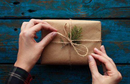 A woman's hands tying a bow in twine around a small brown package with a single twig of evergreen tucked just beneath the twine as  a decorative element
