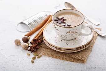 A white table with some fabric and ingredients for clove chai tea, a grater and a spoon, next to a white cup and saucer with clove chai tea in it