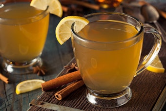 2 Hot Toddy drinks in glass mugs on a wood table with cinnamon sticks and lemons 