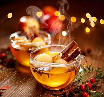 2 clear glass mugs on a wooden table with steaming mulled apple cider - with apples, cloves and cinnamon sticks in it, and small pieces of holiday garlands on the table, and twinkling lights in the background