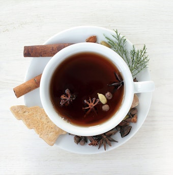 a white cup and saucer filled with clove tea on a white table, with ingredients, a tree-shaped cookie, and a garland on the saucer