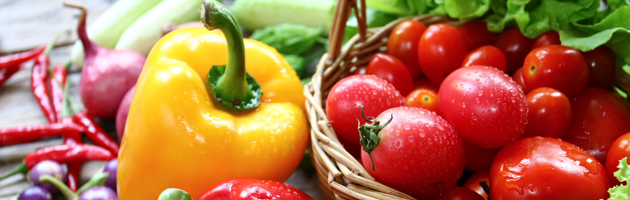 An assortment of hot and bell pepper with shallots in the background next to a basket of cherry and grape tomatoes with lettuce in the background