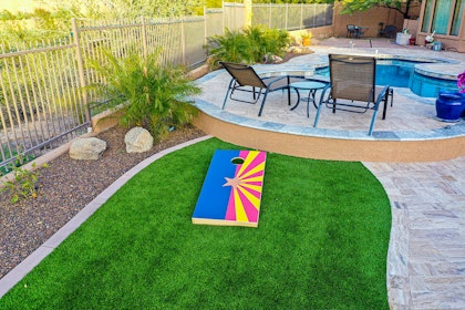 Backyard in Arizona with grass, plants, seating, a pool, and a corn hole board with the AZ Flag printed on it.