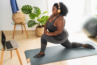 Woman stretching near monstera houseplant, home workout equipment and a laptop.