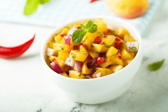 Peach Salsa in a bowl with ingredients nearby on the counter.