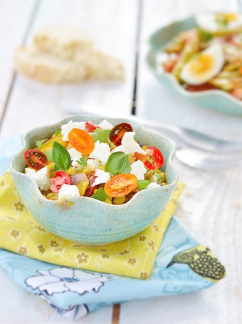 A corn, feta cheese, cherry tomato and basil salad in a bowl near printed napkins on a white wood table.
