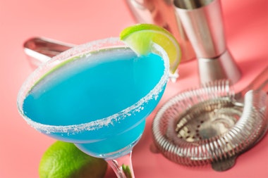 A blue margarita with a lime wedge on a salmon pink table with drink mixing tools and a lime.