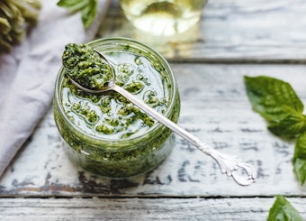 Homemade pesto in a jar on a wooden table with basil and oil nearby.