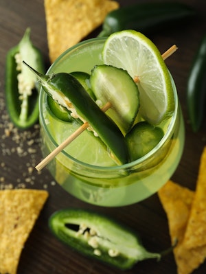 A cucumber jalapeno margarita, on a table with corn chips and jalapeno peppers.