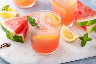 Minty Watermelon Lemonade in a glass on a cutting board with fresh cut lemon, watermelon and mint leaves.