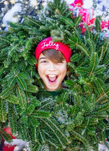 A boy with a red hat on that says jolly on it. He has his head inside a wreath