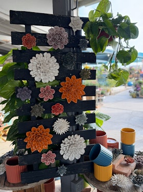 A variety of colorful ceramic flowers hanging on a wood wall display with colorful pots and houseplants nearby.
