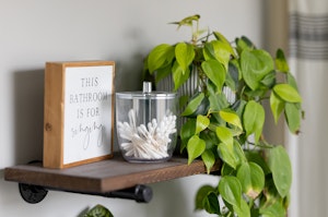 A shelf in a bathroom with a heartleaf philodendron, a glass container with q-tips and a plaque that says,