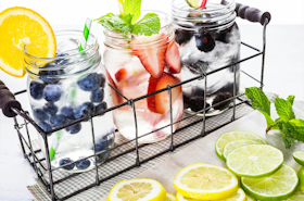 3 water infusions in a wire basket on a table near some sliced lemons, limes and fresh mint - one with blueberries and an orang slice, another with strawberries and mint, and the third with blackberries and lime